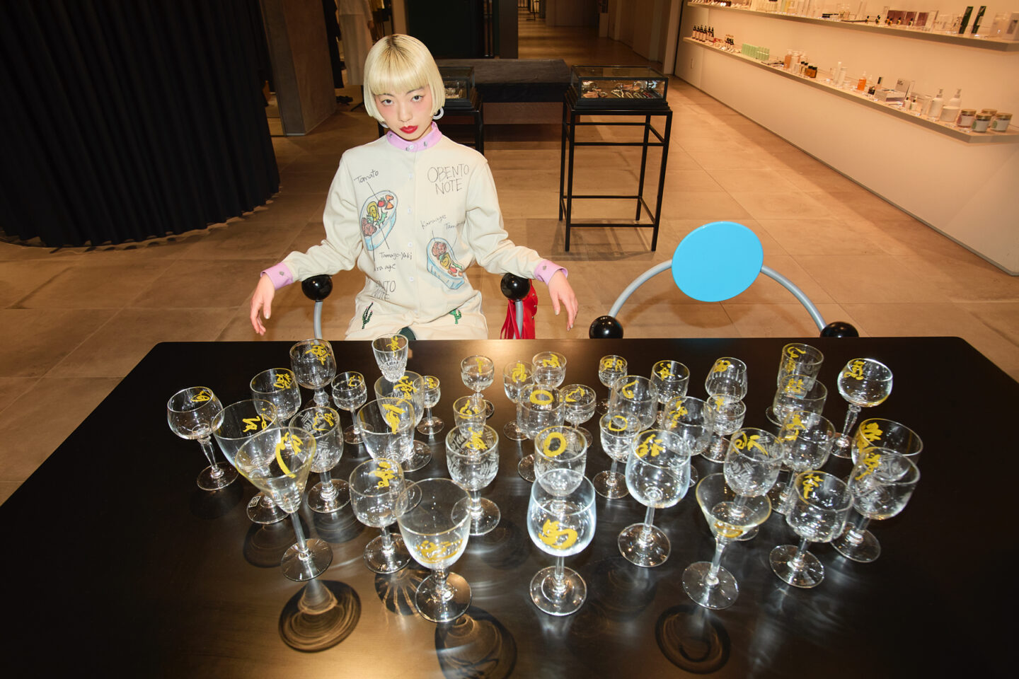 アオイヤマダさんがグラスが並ぶテーブルの前に座るインスタレーション風のシーン